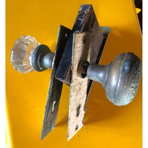 May include: An antique door knob set with a clear glass knob and a tarnished brass knob. The set includes the door plates and hardware, showing signs of age and wear. The hardware is mounted on a yellow surface.