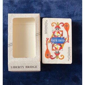 May include: A deck of playing cards with a white box. The card box has the text "LIBERTY BRIDGE" printed on it. The deck of cards shows a joker card with a red and orange design and the text "PLASTIC COATED".