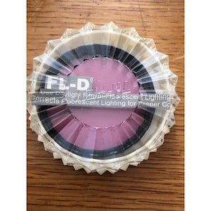 May include: A round, clear plastic filter with a pink center and black and white rings. The filter has a ridged, star-like edge. Text on the filter reads "FL-D" and "Use Daylight Film in Fluorescent Lighting".