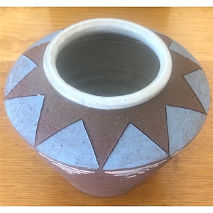Puede incluir: Una olla de cerámica hecha a mano con una boca ancha y un cuerpo redondeado. La olla es principalmente marrón con patrones triangulares azul claro alrededor de los lados. El borde es blanco y el interior es de un gris más oscuro.