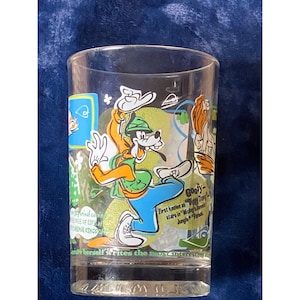 May include: A clear glass featuring a colorful cartoon illustration of Goofy, with text that reads "Goofy - First known as 'Dippy Dawg' stars in 'Mickey's Jungle Parade'". The glass also has other cartoon characters and text.