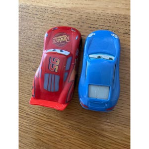 May include: Two toy cars, one red and one blue. The red car features the number 95, Rust-eze branding, and cartoon eyes on the windscreen. The blue car has a smooth finish and cartoon eyes on the windscreen. Both vehicles are small scale models.