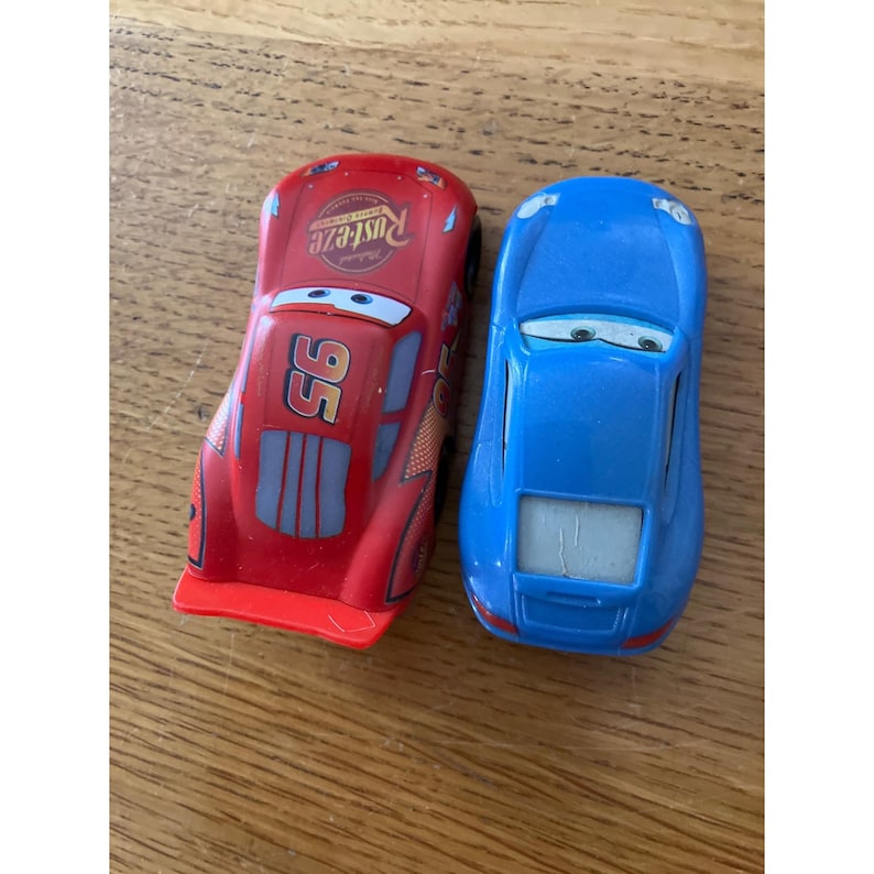 May include: Two toy cars, one red and one blue. The red car features the number 95, Rust-eze branding, and cartoon eyes on the windscreen. The blue car has a smooth finish and cartoon eyes on the windscreen. Both vehicles are small scale models.