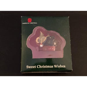 May include: A dark green rectangular box featuring the American Greetings logo and the text Sweet Christmas Wishes. Inside a die-cut window, a small figurine depicts a grey mouse and a black cat resting together on a red base with text.