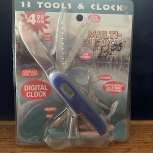 May include: A blue multi-purpose tool with a digital clock embedded in the handle, packaged on a card labeled 13 TOOLS & CLOCK. The tool features a stainless steel knife, wood saw, can opener, scissors, bottle opener, flathead screwdriver, reamer, and file.