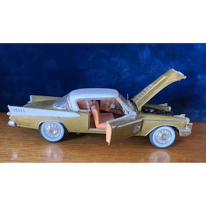 Könnte beinhalten: Ein gold-weißes Vintage-Modellauto mit geöffneter Motorhaube und Tür. Das Auto hat Weißwandreifen und ein weißes Dach. Der Innenraum ist orange. Das Auto steht auf einer Holzoberfläche.