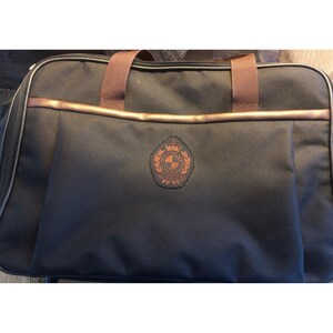 Puede incluir: Bolso de viaje negro con detalles marrones. Cuenta con un asa marrón y un emblema decorativo en la parte delantera. El bolso es rectangular con cierre de cremallera. El ribete y el emblema son de color marrón claro.