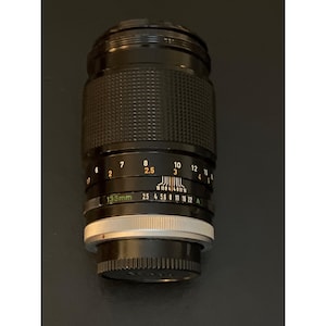 Puede incluir: Un objetivo de c&aacute;mara negro de 135 mm con un anillo de enfoque texturizado, una escala de profundidad de campo y un anillo de apertura con marcas de 2,5 a 22. El objetivo incluye un anillo de montaje plateado y una tapa trasera negra.