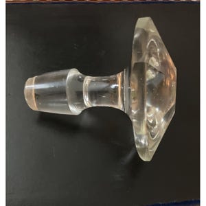 May include: A clear, faceted glass bottle stopper. The stopper has a cylindrical stem and a geometric, diamond-shaped top. The glass is transparent, allowing light to pass through. The stopper is likely for a wine or liquor bottle.