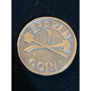 May include: A circular metallic coin with a gold-toned finish. The word BRAZEN is embossed in an arc along the top edge, and the word COIN is embossed along the bottom edge. In the center, two crossed hammers are depicted in relief.