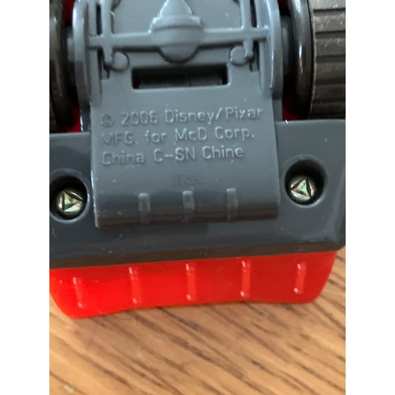 May include: The underside of a red toy vehicle featuring a grey plastic chassis with two visible triangular screws. Embossed text reads: 2006 Disney/Pixar MFG. for McD Corp. China C-SN Chine. The toy has black wheels and a bright red body section.