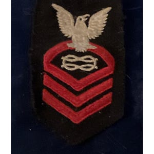 May include: A black patch featuring a white eagle at the top, a red emblem in the middle, and three red chevrons at the bottom. The patch is likely a military insignia or rank identifier.