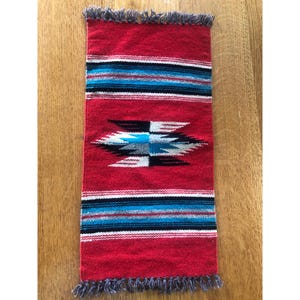 May include: A rectangular, woven textile in red with a geometric pattern. The design features horizontal stripes in black, white, and blue, and a central motif with white, black, and blue elements. The edges have a gray fringe.