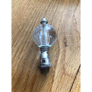 Op de afbeelding: Transparante glazen finial met een geribbelde, bolvormige globe en een zilverkleurige metalen basis. De finial heeft een kleine, afgeronde top en een schroefdraadbasis. Waarschijnlijk voor een lamp of decoratief item.