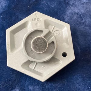 May include: A light gray hexagonal component featuring a central circular metallic insert. The piece includes molded markings, including the word LENS with an upward-pointing arrow, the letter D, and the number 5, along with a small circular hole.