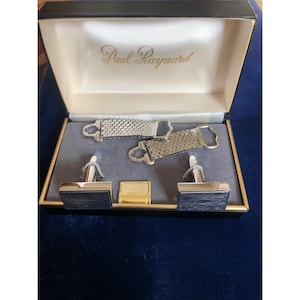 May include: A vintage Paul Raynard cufflink and tie clip set in a black box. The set includes two silver-toned tie clips, two cufflinks with dark gray rectangular faces, and a gold-toned tie bar. The box has a cream-colored interior.