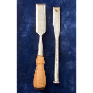 May include: Two wood chisels on a dark blue background. One chisel has a wooden handle and a silver blade. The other chisel is all silver. The wooden handle chisel has the text "USA" on the blade.