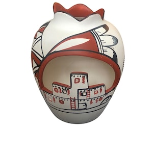 May include: A white ceramic vase with a scalloped rim, featuring a large cutout opening. The exterior is decorated with hand-painted geometric patterns in red and black, and a detailed illustration of a multi-level pueblo dwelling.