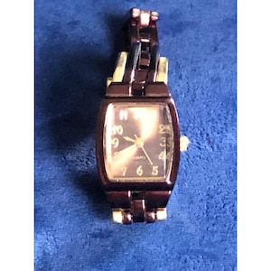 May include: A rectangular brown quartz watch with a gold-tone face and hands. The watch has a brown metal band with gold-tone accents. The watch face displays the numbers 12, 3, 6, and 9.