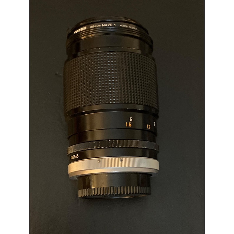 Puede incluir: Un objetivo de c&aacute;mara negro con anillo de enfoque texturizado, marcas de distancia en naranja en pies y metros, y montura de metal plateado. El elemento frontal indica 58mm Haze 1 y Made in U.S.A. El cuerpo incluye el n&uacute;mero de serie 135545.