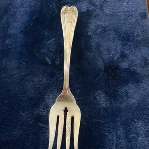 May include: A silver-toned metal fork featuring a three-pronged design with a central cutout. The handle is elongated and tapers toward the top, ending in a decorative scroll-like motif with an engraved monogram consisting of three letters.