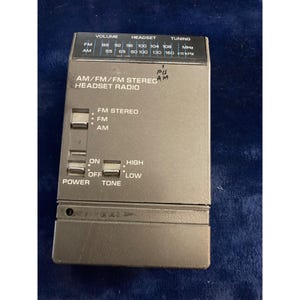 May include: A gray AM/FM/FM stereo headset radio. The radio has buttons for power, tone, and FM stereo. It also has a volume and tuning dial. The radio has the text "AM/FM/FM STEREO HEADSET RADIO" printed on it.