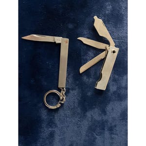 Puede incluir: Dos navajas multiusos de color plateado con acabados metálicos. Una herramienta presenta una hoja extendida y una anilla circular para llaves en la base. La segunda herramienta está parcialmente desplegada, mostrando tres utensilios metálicos.