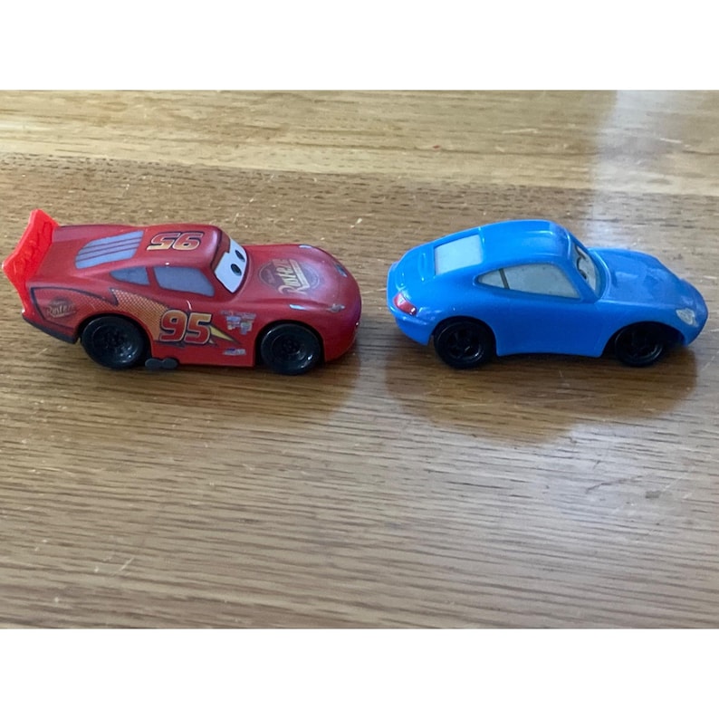 May include: Two small toy cars, one red and one blue. The red car features the number 95, yellow lightning bolt graphics, and a rear spoiler. The blue car is a smooth, rounded coupe model. Both vehicles have black wheels and white windshields.
