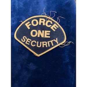May include: A navy blue patch with the words "FORCE ONE SECURITY" in gold lettering. The patch has a gold border and is in a shield shape. The patch is on a dark blue background.