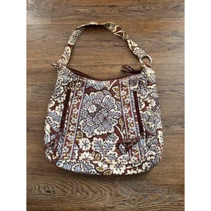 Puede incluir: Bolso de hombro con estampado floral en tonos marrón, azul y crema. El bolso presenta un gran diseño floral central y una correa a juego. Tiene cierre de cremallera y bolsillos exteriores.