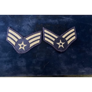 May include: Two blue and beige Air Force rank insignia patches. Each patch features a star within a circle and three horizontal stripes. The patches are displayed on a dark blue surface.