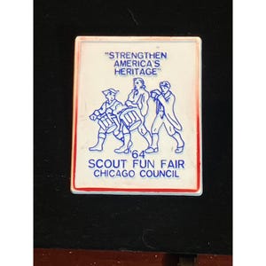 May include: A rectangular white pin with a red border. The pin features blue line art of three figures playing drums and a banner that reads "STRENGTHEN AMERICA'S HERITAGE". Below the figures is the text "'64 SCOUT FUN FAIR CHICAGO COUNCIL".
