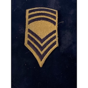 Op de afbeelding: Een vintage militaire patch met een gouden achtergrond en donkerblauwe strepen. De patch heeft drie gebogen strepen aan de bovenkant en drie chevronstrepen aan de onderkant, allemaal omlijnd in donkerblauw. De patch is waarschijnlijk voor een militaire rang.