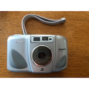 May include: Silver Kodak Advantix C750 camera with a gray wrist strap. The camera has a lens with the text "Kodak Aspheric Zoom Lens 2.5x" and the Kodak logo. The camera is on a wooden surface.