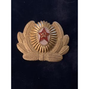 May include: A gold-colored military hat badge with a red star and hammer and sickle emblem at the center. The badge features a wreath design with detailed feather-like elements and a sunburst pattern.