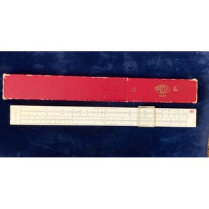 May include: A vintage slide rule with a red case. The slide rule is off-white with black markings and a clear cursor. The red case has the word "POST" and the number "1447" printed on it. A star is also visible on the case.