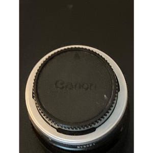 Puede incluir: Una tapa de objetivo de c&aacute;mara Canon circular y negra con un borde estriado y texturizado, que presenta el logotipo de Canon en relieve en el centro y una peque&ntilde;a marca triangular de alineaci&oacute;n en la parte superior, sobre un fondo oscuro.
