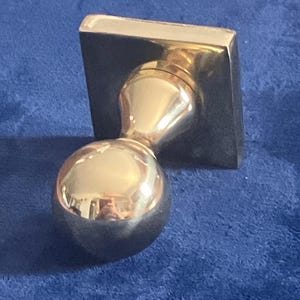 May include: A polished gold-tone metal door knob featuring a spherical handle attached to a square-shaped backplate. The reflective surface of the sphere shows a distorted view of the surroundings, and the hardware has a smooth, metallic finish.