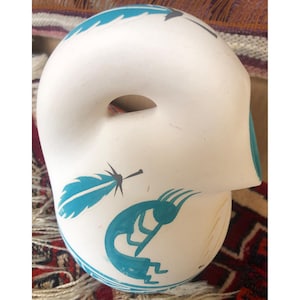 Op de afbeelding: Een wit keramisch masker met een grote opening en een gebogen vorm. Het masker is versierd met turquoise veren en een Kokopelli-figuur. Het masker ligt op een patroontextiel.