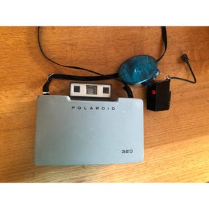 May include: A vintage Polaroid 320 instant camera in a light blue-gray color. The camera has the word "POLAROID" printed on the front, along with the number "320". A black strap, flash, and flash attachment are also visible.