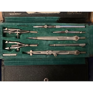 以下が含まれることがあります： 緑色のベルベットの内装が施された黒いケースには、ヴィンテージの製図道具一式が収められています。このセットには、コンパス、ディバイダー、その他の精密機器が含まれており、すべて銀色の金属製です。ケースには「ACADEMIC」と「SCRANTON, PA MADE IN GERMANY」と記載されています。