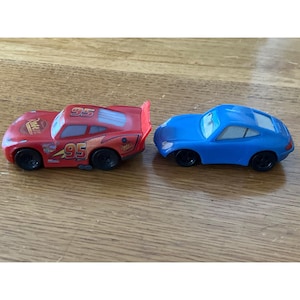 May include: Two toy cars, one red and one blue. The red car features the number 95, Rust-eze branding, and a rear spoiler. The blue car is a solid colour with black wheels and white-rimmed eyes on the windscreen. Both vehicles have black wheels.
