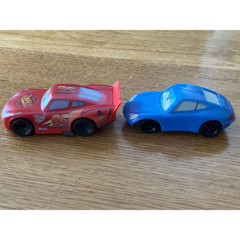 May include: Two toy cars, one red and one blue. The red car features the number 95, Rust-eze branding, and a rear spoiler. The blue car is a solid colour with black wheels and white-rimmed eyes on the windscreen. Both vehicles have black wheels.