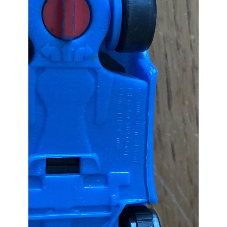 May include: The underside of a blue toy vehicle featuring black wheels and a circular red component. Embossed text on the plastic chassis includes manufacturing details and copyright information, with directional arrows visible near the red part.