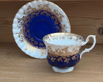 Op de afbeelding: Een vintage theekop en schotel set. De kop en schotel hebben een wit en goud bloemmotief met een diepblauwe band. De theekop heeft een gebogen oor en staat op een kleine voet. De schotel heeft een bijpassend ontwerp.