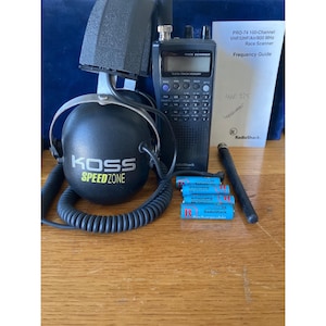 Puede incluir: Un escáner de carreras RadioShack negro con una guía de frecuencias, auriculares y baterías recargables. Los auriculares son negros con el texto "KOSS SPEEDZONE" en verde y blanco. El escáner tiene una pantalla digital y varios botones.