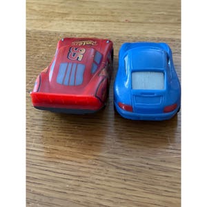 May include: A red toy race car with the number 95 and Rust-eze branding on the roof, positioned next to a blue toy sports car. Both vehicles are viewed from the rear, showing their respective spoilers, taillights, and rear window designs.