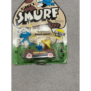 Könnte beinhalten: Ein Vintage Smurf-about Spielzeugauto aus Druckgussmetall. Das braune Auto hat eine blaue Schlümpfe-Figur mit gelbem Hut. Die Verpackung enthält das Wort "SMURF" und den Text "Redwood Smurf-about".