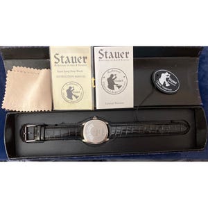 May include: A Stauer jump hour watch with a black crocodile-embossed leather strap and silver-tone case, presented in a black box with an instruction manual, a warranty booklet, a cleaning cloth, and a round black hang tag with white text.