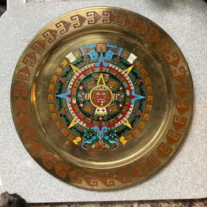 May include: A round, gold-colored metal plate with a detailed, colorful central design. The design features a sun-like face surrounded by geometric shapes in red, blue, green, and yellow. The outer rim has a repeating brown pattern.